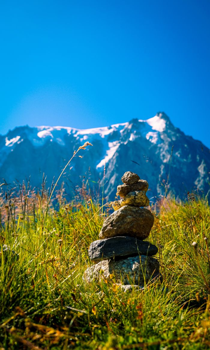 Stacked rocks on grassy field with snow-capped mountain in background, bright day.